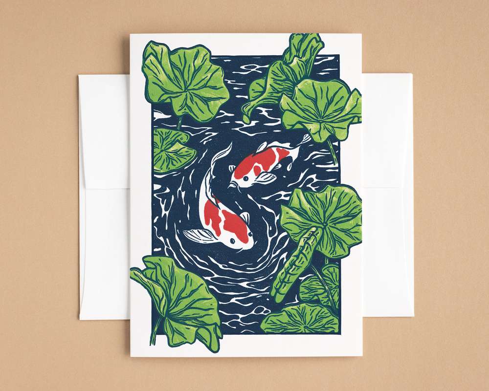 A vertical white card blue pond with green lily pads and white/red koi fish. The card sits on top of a white envelope, which lies on top of a brown backdrop.